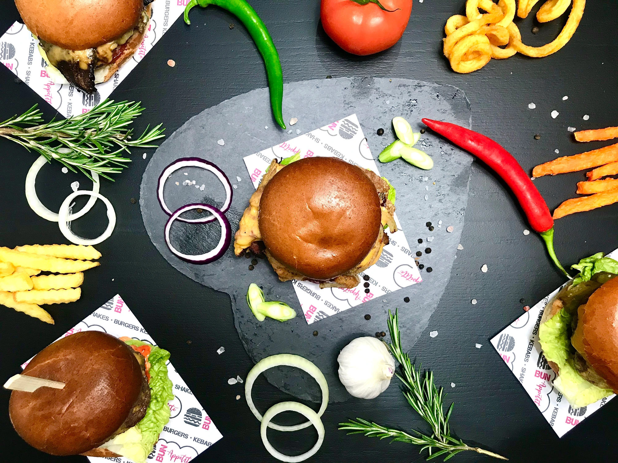 Burgers, fries and fresh ingredients arranged on slate with Bun Appetite branded paper